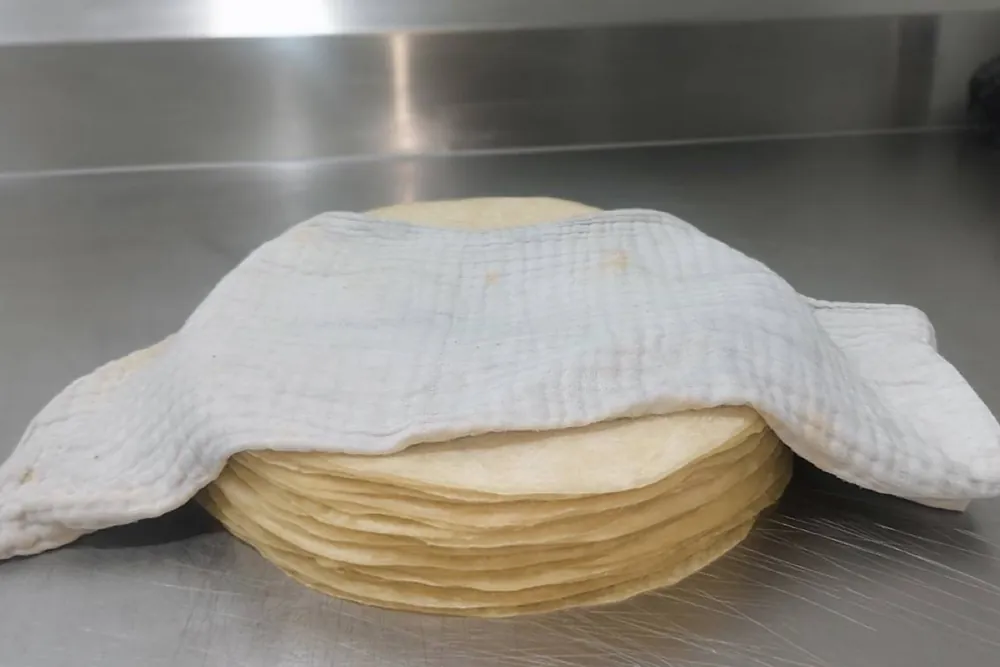 A stack of Sunrise Tacos flour tortillas covered with a cloth towel on a steel counter