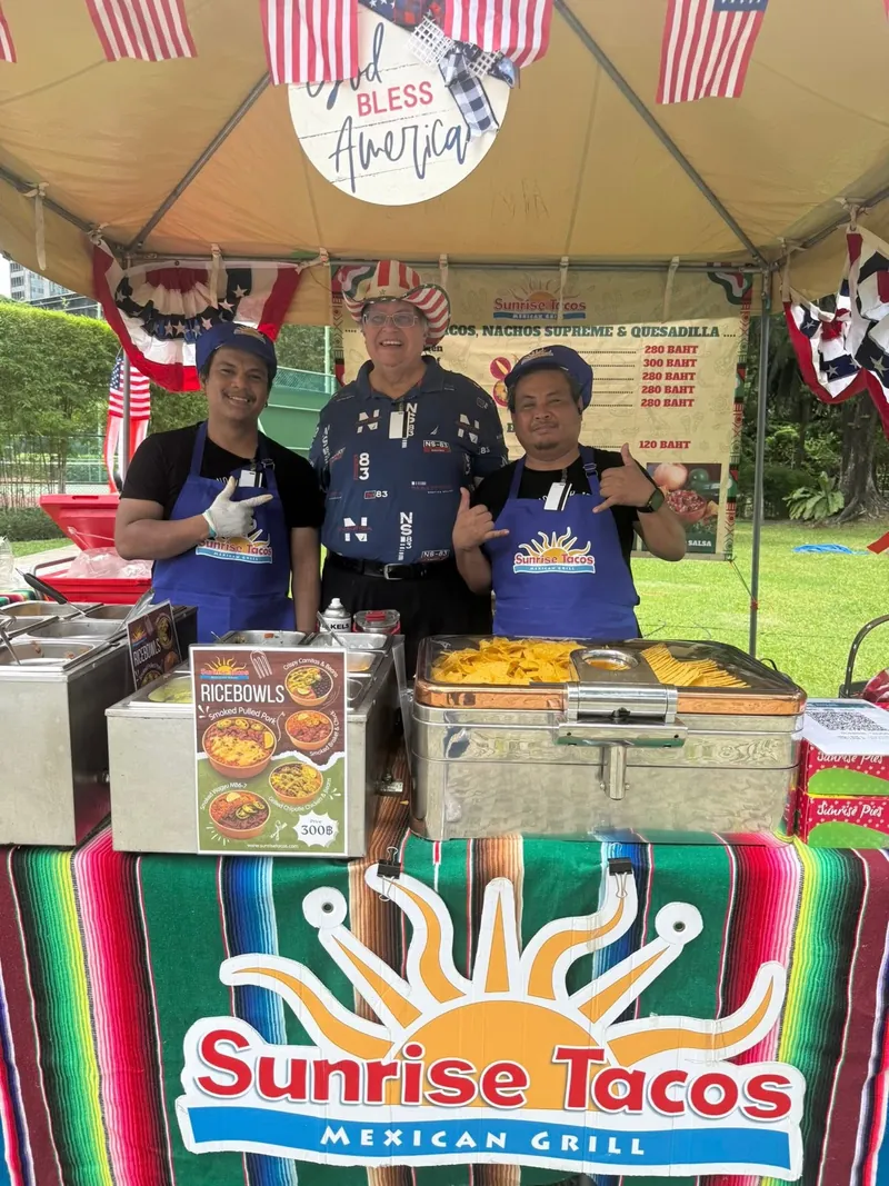Greg Lange in a stars-and-stripes hat with two Sunrise Tacos team members in blue aprons at an event stall behind a large Sunrise Tacos Mexican Grill cloth